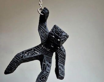 May include: A black, 3D-printed keychain pendant in the shape of a stylized human figure. The pendant is suspended from a silver chain. The figure has an intricate, geometric pattern throughout its form.