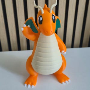 May include: A small orange dragon figurine with a cream-colored belly, teal inner wings, and two long antennae on its head. The creature has large eyes, white claws on its feet and hands, and is posed with one arm raised in a waving gesture.