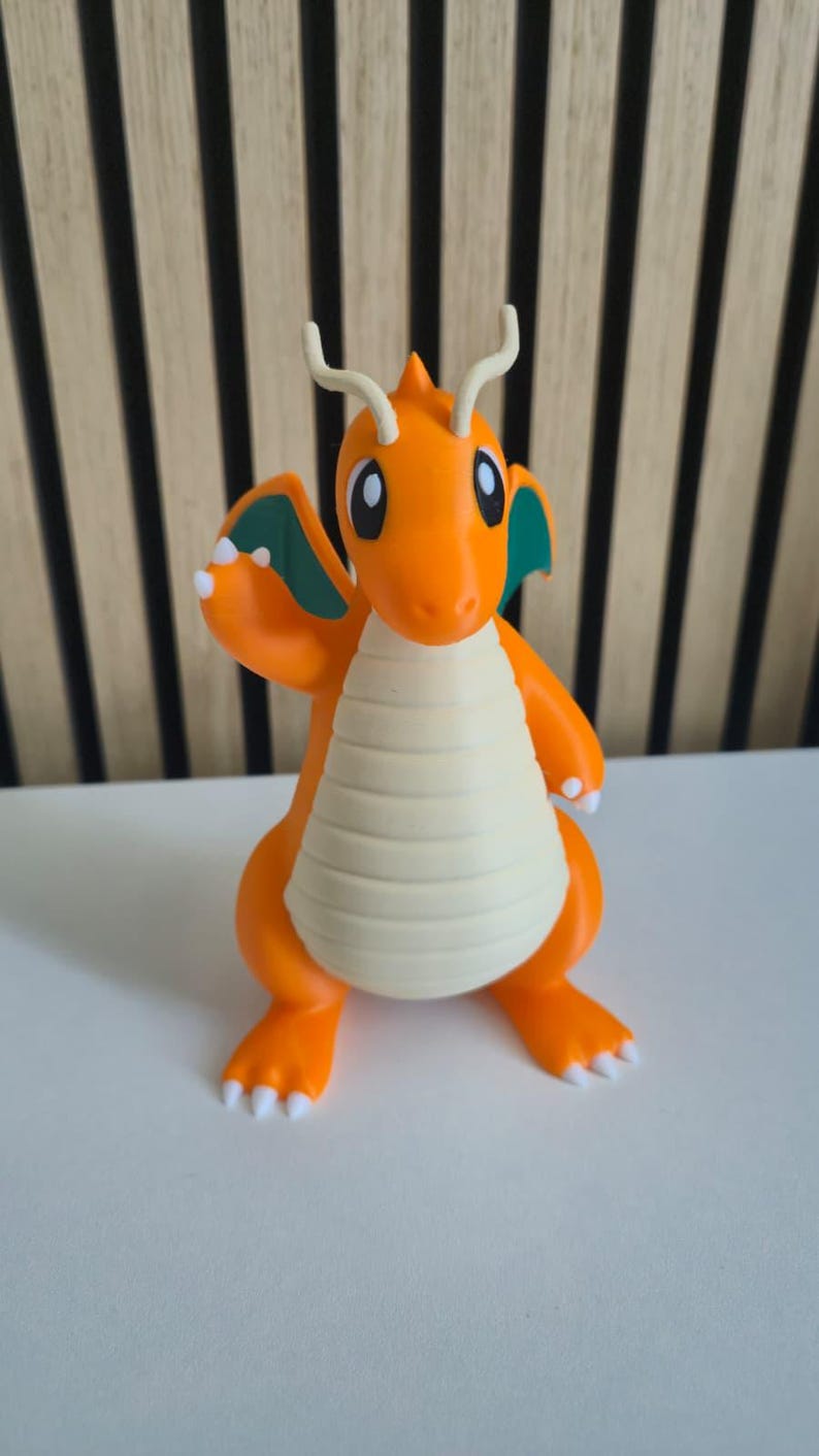 May include: A small orange dragon figurine with a cream-colored belly, teal inner wings, and two long antennae on its head. The creature has large eyes, white claws on its feet and hands, and is posed with one arm raised in a waving gesture.