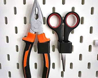 Könnte beinhalten: Eine orangefarbene und schwarze Zange und eine schwarze Schere mit roten Akzenten sind auf einer weißen Lochwand montiert. Die Werkzeuge werden von schwarzen Haltern gehalten. Die Lochwand hat gleichmäßig verteilte Löcher.