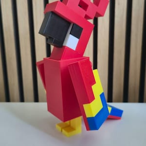 May include: A blocky, pixelated parrot figurine constructed from red, yellow, and blue plastic components. The bird features a prominent black beak, a red crest, and a square-shaped body design, standing on two small yellow feet on a flat surface.