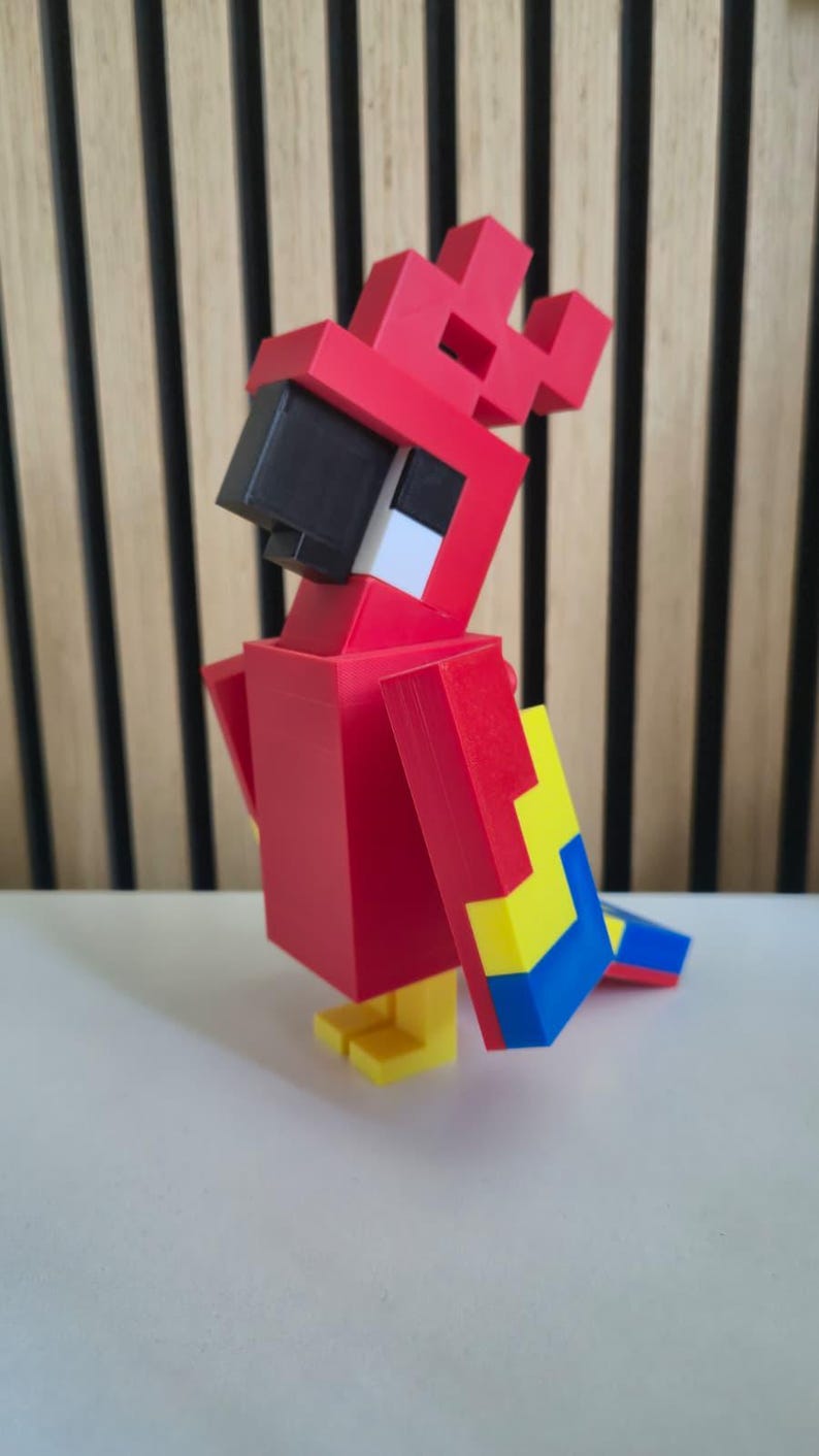 May include: A blocky, pixelated parrot figurine constructed from red, yellow, and blue plastic components. The bird features a prominent black beak, a red crest, and a square-shaped body design, standing on two small yellow feet on a flat surface.