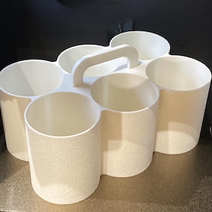 May include: A white plastic caddy featuring six cylindrical compartments arranged in two rows of three. The organizer includes a central integrated handle for carrying and has a textured exterior finish on the outer walls of the circular containers.