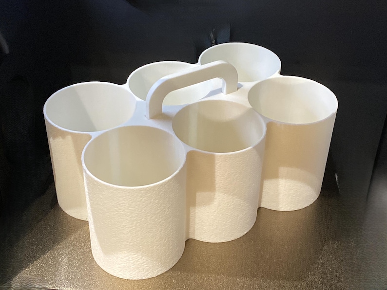 May include: A white plastic caddy featuring six cylindrical compartments arranged in two rows of three. The organizer includes a central integrated handle for carrying and has a textured exterior finish on the outer walls of the circular containers.