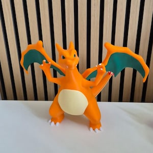 May include: A bright orange dragon-like figurine with a cream-colored belly, teal-colored inner wings, and white claws. The creature stands on two legs with its wings spread wide, featuring a long tail and a sharp, pointed head with small horns.