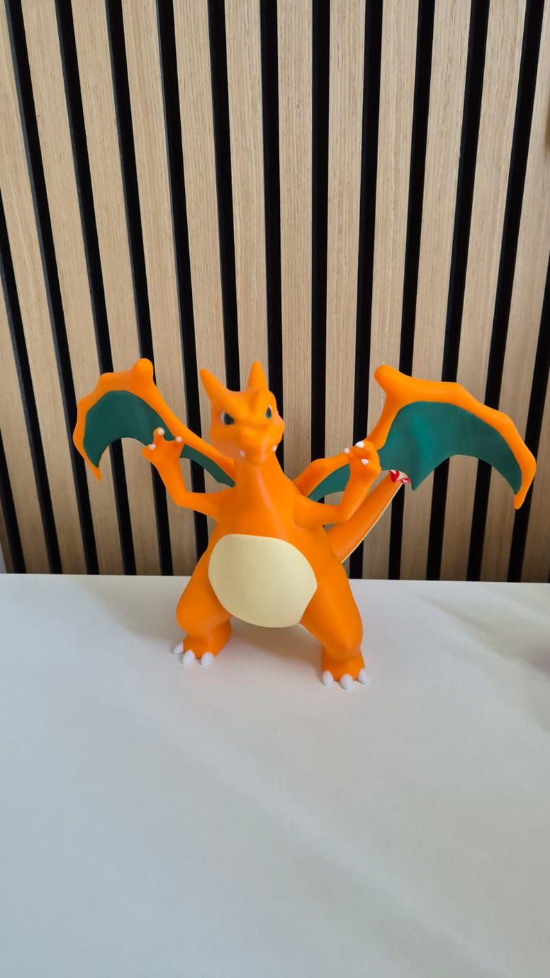 May include: A bright orange dragon-like figurine with a cream-colored belly, teal-colored inner wings, and white claws. The creature stands on two legs with its wings spread wide, featuring a long tail and a sharp, pointed head with small horns.