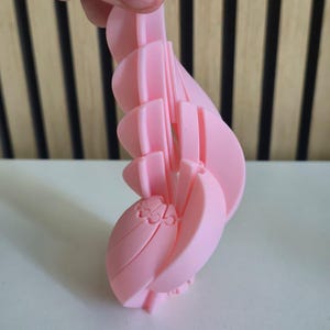 May include: A light pink, 3D-printed articulated object featuring a series of curved, interlocking segments that form a spiral or wing-like structure. The surface has a smooth matte finish, and the design consists of multiple connected pieces.