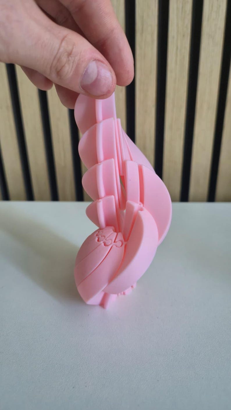May include: A light pink, 3D-printed articulated object featuring a series of curved, interlocking segments that form a spiral or wing-like structure. The surface has a smooth matte finish, and the design consists of multiple connected pieces.