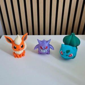 May include: Three small plastic character figurines arranged in a row. On the left is an orange and cream fox-like creature. In the center is a purple, spiky-haired creature with red eyes. On the right is a light blue creature with a green bulb.