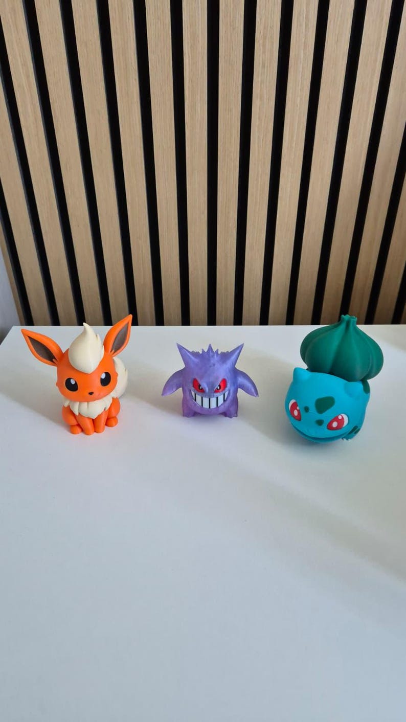 May include: Three small plastic character figurines arranged in a row. On the left is an orange and cream fox-like creature. In the center is a purple, spiky-haired creature with red eyes. On the right is a light blue creature with a green bulb.