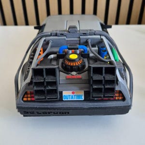 May include: A gray scale model of a sports car featuring a rear engine compartment with blue, orange, and green accents. The vehicle has black exhaust vents, a license plate that reads OUTATIME, and the word DeLorean embossed on the rear bumper.