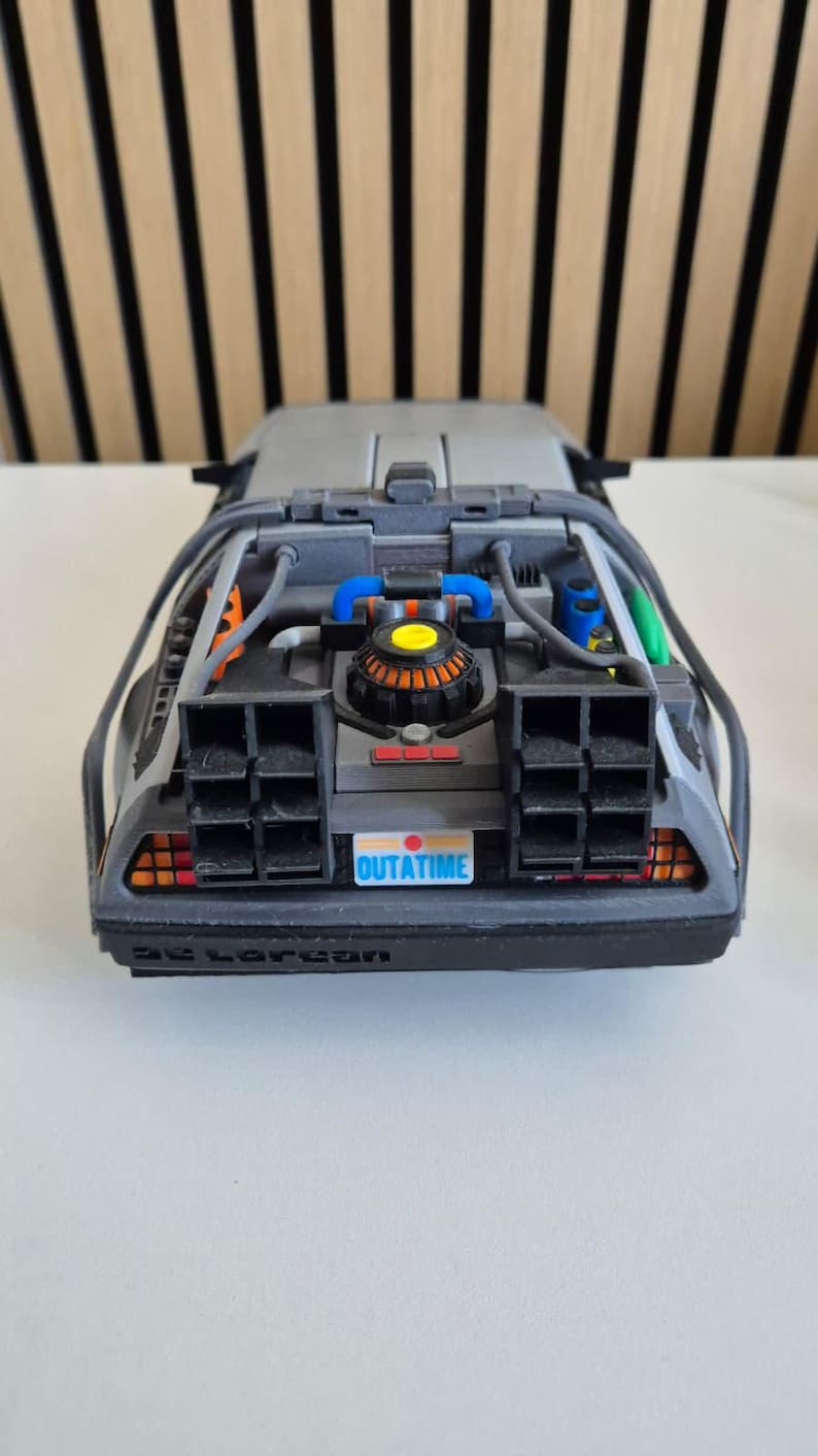 May include: A gray scale model of a sports car featuring a rear engine compartment with blue, orange, and green accents. The vehicle has black exhaust vents, a license plate that reads OUTATIME, and the word DeLorean embossed on the rear bumper.