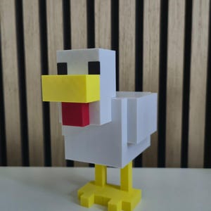 May include: A blocky, pixelated chicken figurine constructed from white, yellow, and red plastic components. The figure features a white body and head, a prominent yellow beak, a red wattle, black square eyes, and yellow feet with three toes.