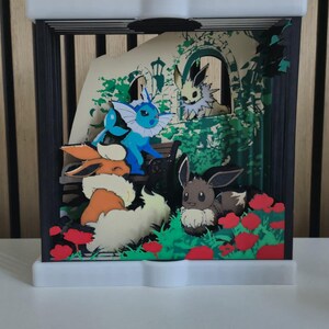 May include: A multi-layered shadow box display featuring several Eevee evolution characters including Vaporeon, Flareon, and Eevee in a garden setting with red flowers, green foliage, and a stone archway, all enclosed in a black and white frame.