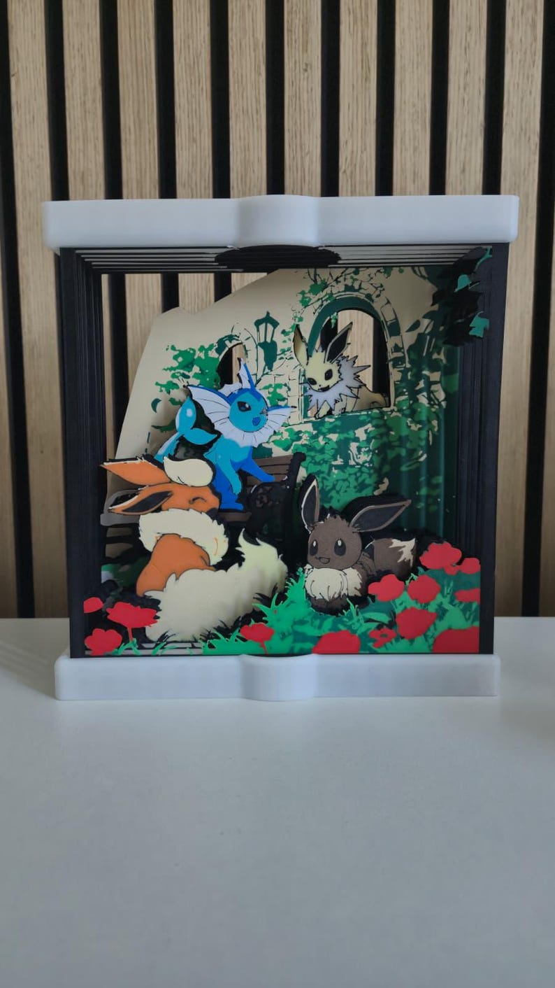 May include: A multi-layered shadow box display featuring several Eevee evolution characters including Vaporeon, Flareon, and Eevee in a garden setting with red flowers, green foliage, and a stone archway, all enclosed in a black and white frame.