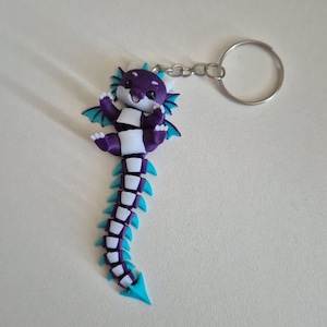 May include: A purple and white articulated dragon keychain with bright teal accents on the wings, fins, and tail tip. The dragon has a smiling face, small white claws, and is attached to a circular silver metal split ring by a short chain.
