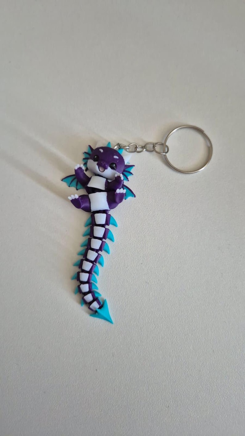 May include: A purple and white articulated dragon keychain with bright teal accents on the wings, fins, and tail tip. The dragon has a smiling face, small white claws, and is attached to a circular silver metal split ring by a short chain.