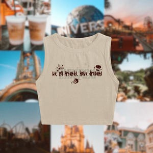 Puede incluir: Un top corto beige con cuello redondo. El top presenta el texto "We, my friends, have dragons" en color marrón oscuro, junto con pequeños iconos decorativos. El top es sin mangas.