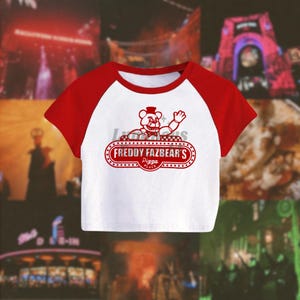 May include: White crop top with red sleeves and collar. The shirt features a red graphic of a bear wearing a hat and waving, with the text "FREDDY FAZBEAR'S Pizza Place" in a red oval design. The shirt is a raglan style.