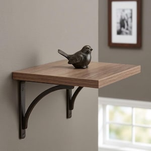 May include: A small, decorative bronze bird figurine sits on a square, wooden floating shelf. The shelf is supported by two dark metal brackets attached to a neutral-colored wall. A framed black and white photo hangs in the background.