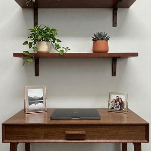 May include: A wooden desk with a laptop, two framed photos, and a drawer. Two shelves above hold plants in pots and a small stack of books. The desk and shelves are made of dark wood, creating a warm, inviting workspace.