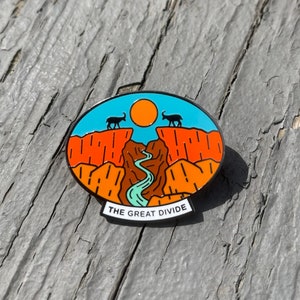 May include: A blue and orange enamel pin with a mountain range and a river. The pin has a white banner at the bottom that reads "THE GREAT DIVIDE".