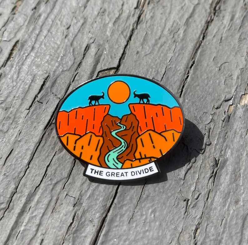 May include: A blue and orange enamel pin with a mountain range and a river. The pin has a white banner at the bottom that reads "THE GREAT DIVIDE".