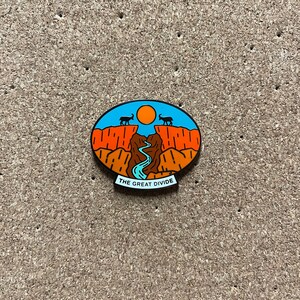 May include: A blue and orange enamel pin with a mountain range and a river, with the text "THE GREAT DIVIDE" at the bottom.
