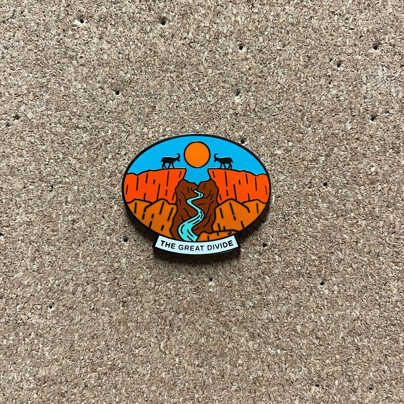 May include: A blue and orange enamel pin with a mountain range and a river, with the text "THE GREAT DIVIDE" at the bottom.