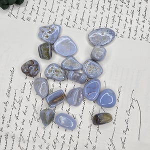 Puede incluir: Una colecci&oacute;n de piedras de &aacute;gata de encaje azul pulidas con patrones blancos y marrones. Las piedras est&aacute;n dispuestas sobre un fondo de papel vintage con escritura manuscrita y una ramita de vegetaci&oacute;n.