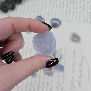 Puede incluir: Una piedra preciosa pulida de color azul claro se sostiene entre los dedos, con otras dispersas cerca. La piedra tiene una forma suave y redondeada y una apariencia nublada. El fondo incluye escritura manuscrita y vegetaci&oacute;n.