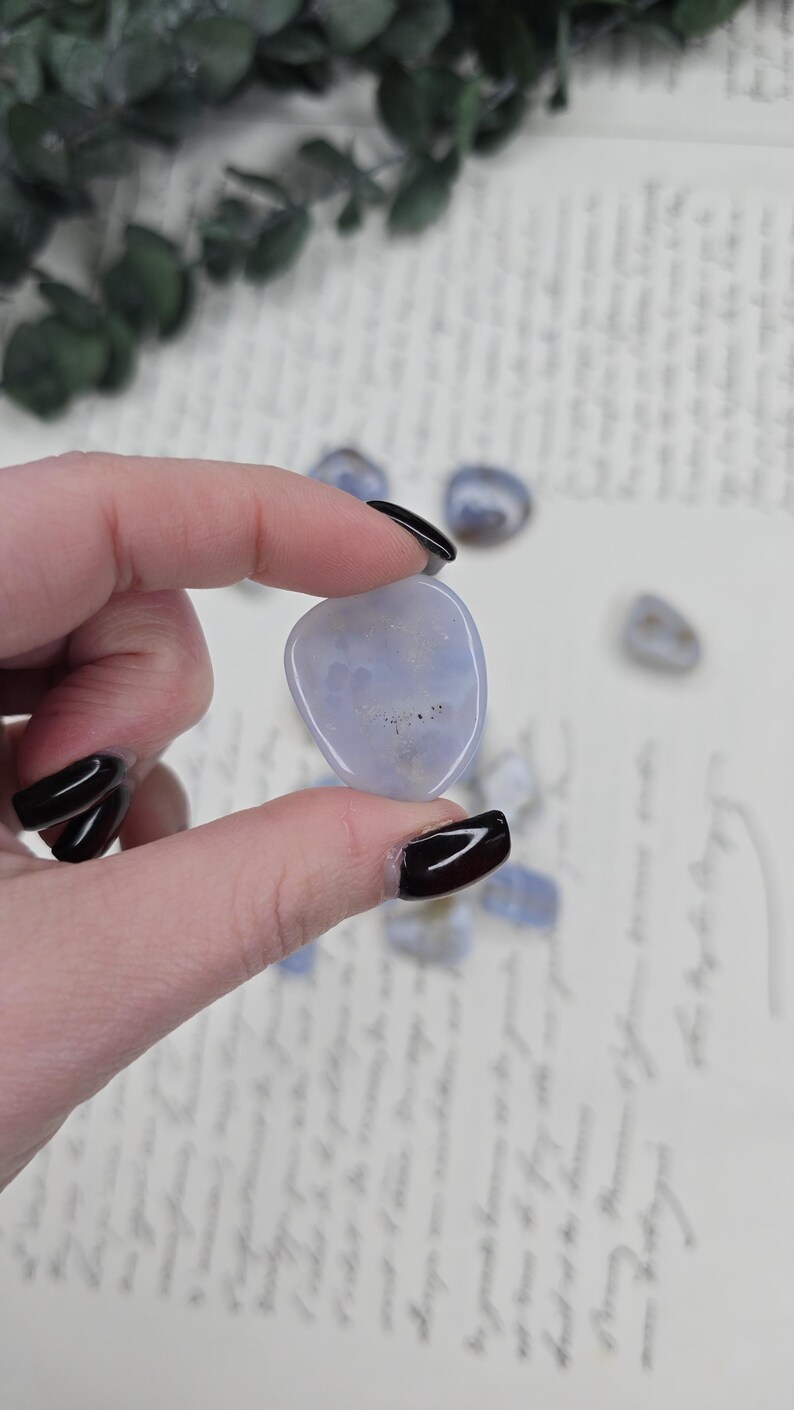 Puede incluir: Una piedra preciosa pulida de color azul claro se sostiene entre los dedos, con otras dispersas cerca. La piedra tiene una forma suave y redondeada y una apariencia nublada. El fondo incluye escritura manuscrita y vegetaci&oacute;n.