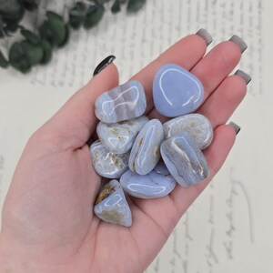 Puede incluir: Un pu&ntilde;ado de piedras de &aacute;gata de encaje azul pulidas. Las piedras son de color azul claro con vetas blancas y marrones. Las piedras son lisas y redondeadas, y var&iacute;an en tama&ntilde;o. El fondo es una imagen borrosa de un documento manuscrito.