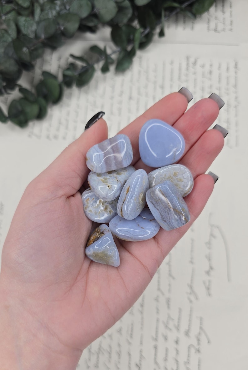 Puede incluir: Un pu&ntilde;ado de piedras de &aacute;gata de encaje azul pulidas. Las piedras son de color azul claro con vetas blancas y marrones. Las piedras son lisas y redondeadas, y var&iacute;an en tama&ntilde;o. El fondo es una imagen borrosa de un documento manuscrito.