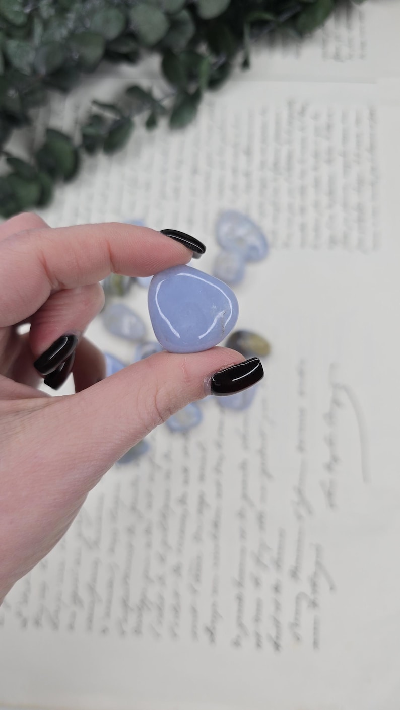 Puede incluir: Una piedra rodada lisa, de color azul claro, sostenida entre los dedos con esmalte de u&ntilde;as negro. La piedra tiene una forma redondeada y una superficie pulida. Otras piedras similares son visibles en el fondo.