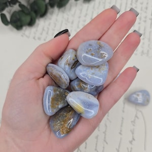 Puede incluir: Un pu&ntilde;ado de piedras de &aacute;gata de encaje azul pulidas. Las piedras tienen un color base azul claro con inclusiones marrones y amarillas. Las piedras lisas y redondeadas se sostienen en una mano abierta, con algunas piedras visibles en el fondo.