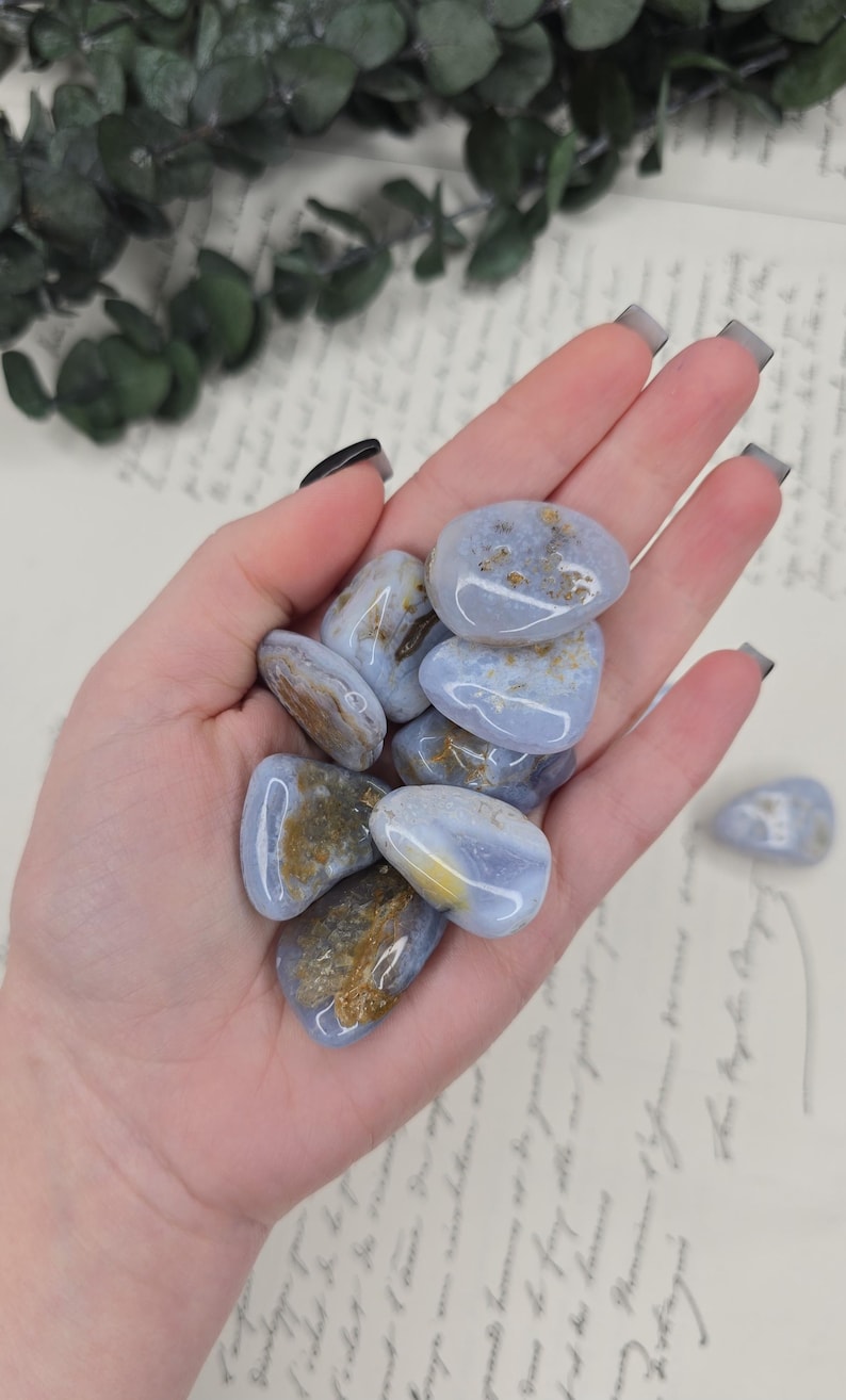 Puede incluir: Un pu&ntilde;ado de piedras de &aacute;gata de encaje azul pulidas. Las piedras tienen un color base azul claro con inclusiones marrones y amarillas. Las piedras lisas y redondeadas se sostienen en una mano abierta, con algunas piedras visibles en el fondo.