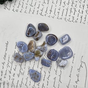 Puede incluir: Una colecci&oacute;n de piedras de &aacute;gata de encaje azul pulidas, cada una con patrones &uacute;nicos y tonos de azul, gris y marr&oacute;n. Estas piedras rodadas se exhiben sobre un documento manuscrito antiguo.