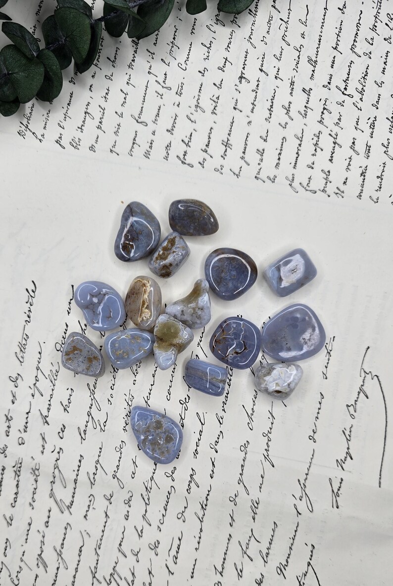 Puede incluir: Una colecci&oacute;n de piedras de &aacute;gata de encaje azul pulidas, cada una con patrones &uacute;nicos y tonos de azul, gris y marr&oacute;n. Estas piedras rodadas se exhiben sobre un documento manuscrito antiguo.
