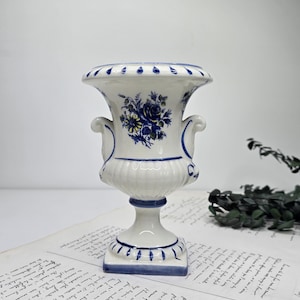 Może przedstawiać: Biała ceramiczna urna z niebieskim wzorem kwiatowym i liściastym. Urna ma dwa uchwyty i podstawę na cokole. Górna krawędź i podstawa są ozdobione niebieskimi akcentami. Urna stoi na kartce papieru z odręcznym tekstem.