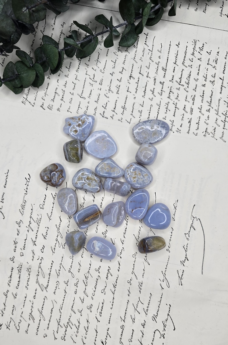 Puede incluir: Una colecci&oacute;n de piedras de &aacute;gata de encaje azul pulidas con patrones blancos y marrones. Las piedras est&aacute;n dispuestas sobre un fondo de papel vintage con escritura manuscrita y una ramita de vegetaci&oacute;n.