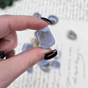 Puede incluir: Una piedra de &aacute;gata azul claro con una superficie pulida, sostenida entre los dedos. La piedra tiene una apariencia nublada con inclusiones marrones y blancas. Otras piedras similares est&aacute;n dispersas en el fondo. El fondo es una superficie blanca con escritura.