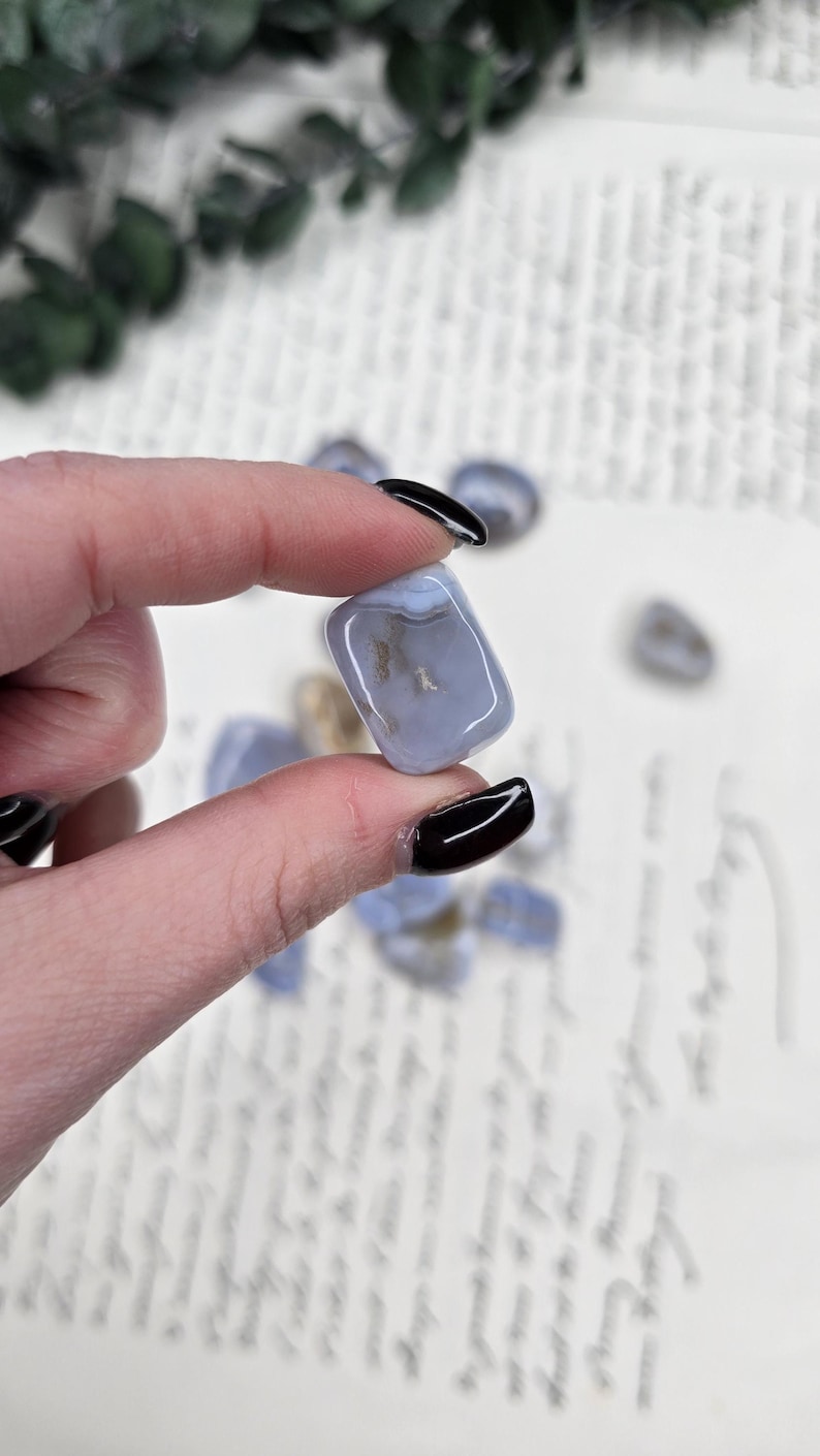 Puede incluir: Una piedra de &aacute;gata azul claro con una superficie pulida, sostenida entre los dedos. La piedra tiene una apariencia nublada con inclusiones marrones y blancas. Otras piedras similares est&aacute;n dispersas en el fondo. El fondo es una superficie blanca con escritura.