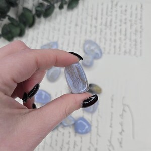 Puede incluir: Una piedra preciosa de &aacute;gata de encaje azul pulida con vetas marrones y blancas se sostiene entre los dedos. Varias otras piedras rodadas est&aacute;n dispersas en el fondo. La &aacute;gata tiene una forma suave y redondeada y es de color azul claro.