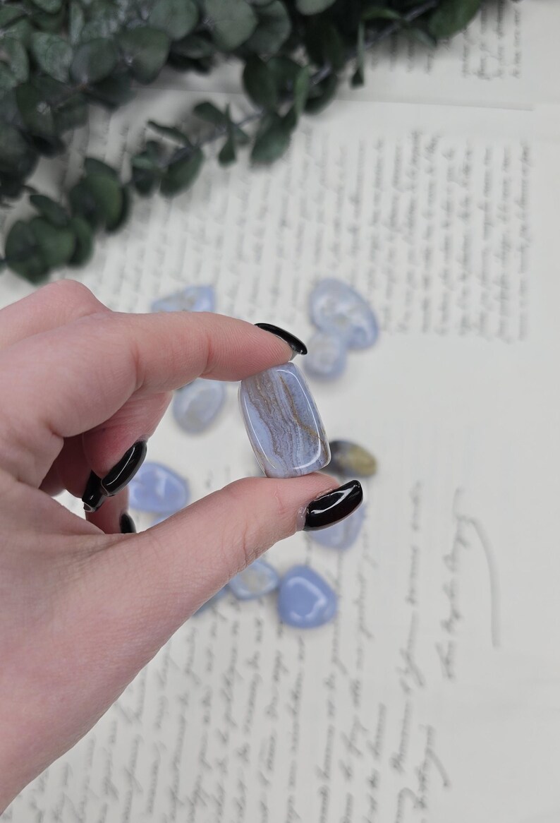 Puede incluir: Una piedra preciosa de &aacute;gata de encaje azul pulida con vetas marrones y blancas se sostiene entre los dedos. Varias otras piedras rodadas est&aacute;n dispersas en el fondo. La &aacute;gata tiene una forma suave y redondeada y es de color azul claro.