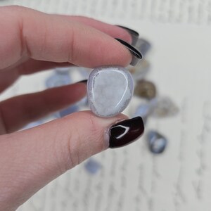 Puede incluir: Una piedra preciosa de &aacute;gata de encaje azul pulida se sostiene entre los dedos. La piedra tiene un color azul p&aacute;lido con inclusiones blancas y transl&uacute;cidas. Otras piedras similares est&aacute;n dispersas en el fondo.