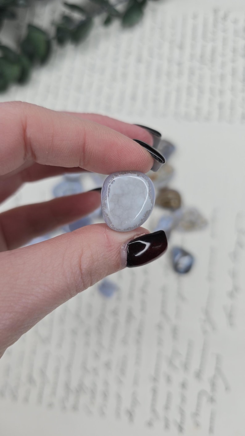 Puede incluir: Una piedra preciosa de &aacute;gata de encaje azul pulida se sostiene entre los dedos. La piedra tiene un color azul p&aacute;lido con inclusiones blancas y transl&uacute;cidas. Otras piedras similares est&aacute;n dispersas en el fondo.