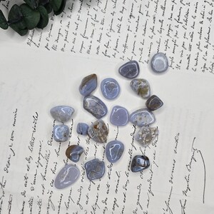 Puede incluir: Una colecci&oacute;n de piedras de &aacute;gata de encaje azul pulidas, cada una con patrones y tonos &uacute;nicos de azul, gris y marr&oacute;n. Las piedras est&aacute;n dispersas sobre un fondo de papel vintage con escritura manuscrita.