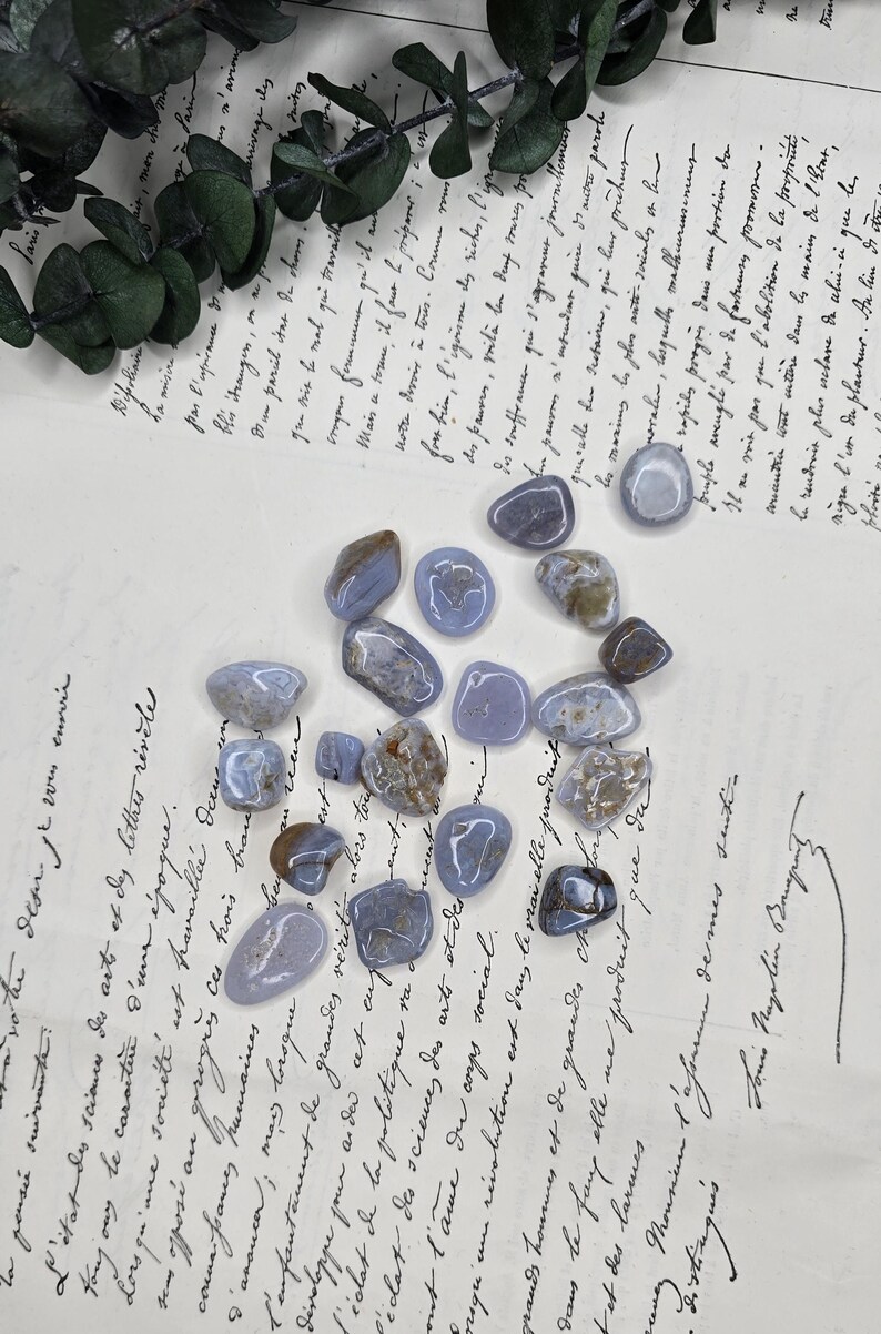 Puede incluir: Una colecci&oacute;n de piedras de &aacute;gata de encaje azul pulidas, cada una con patrones y tonos &uacute;nicos de azul, gris y marr&oacute;n. Las piedras est&aacute;n dispersas sobre un fondo de papel vintage con escritura manuscrita.