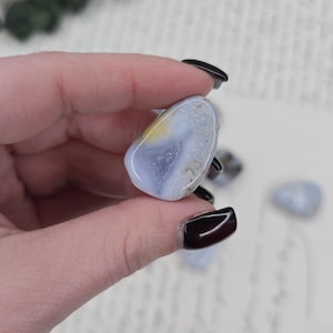 Puede incluir: Una piedra preciosa de &aacute;gata pulida, de color azul, con inclusiones amarillas y blancas. La piedra se sostiene en una mano, con otras piedras similares visibles en el fondo. El &aacute;gata tiene una forma suave y redondeada.